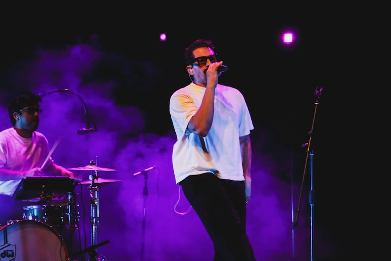 Un hombre canta con micrófono y gafas oscuras mientras otro toca la batería; luces púrpuras crean un ambiente de concierto.