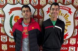 Dos hombres sonrientes en comisaría, uno con chaqueta roja y negro, otro en conjunto deportivo negro con detalles en blanco y verde.