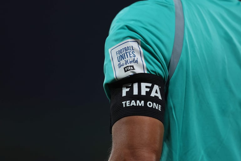 Árbitro de fútbol en camiseta verde y cinta negra que dice 'FIFA TEAM ONE', mientras se prepara en un estadio.