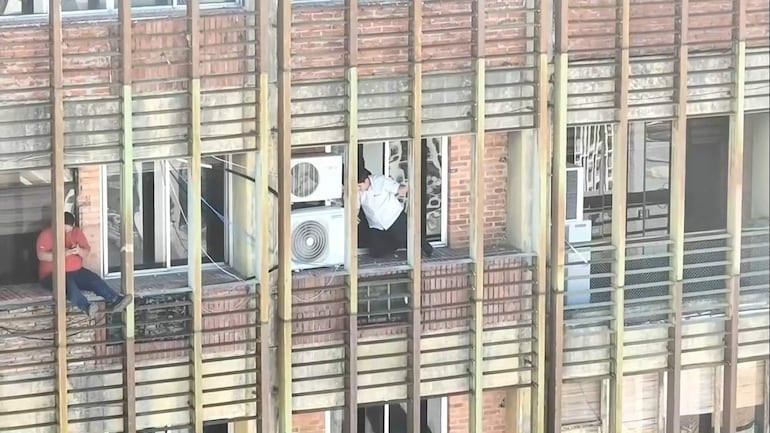 Hombre con camiseta roja sentado en un alféizar y otro con camisa blanca intentando salir por la ventana, rodeados de humo.