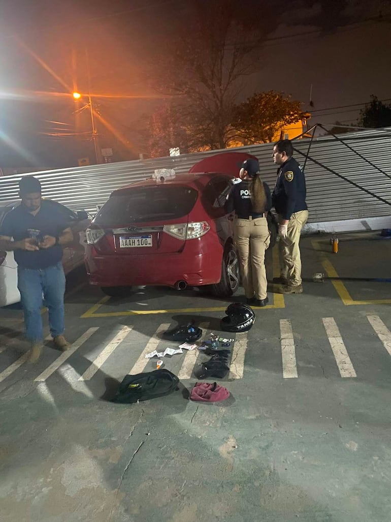 Dos policías en uniforme inspeccionan un auto rojo. Objetos esparcidos en el suelo, ambiente nocturno y tenue.