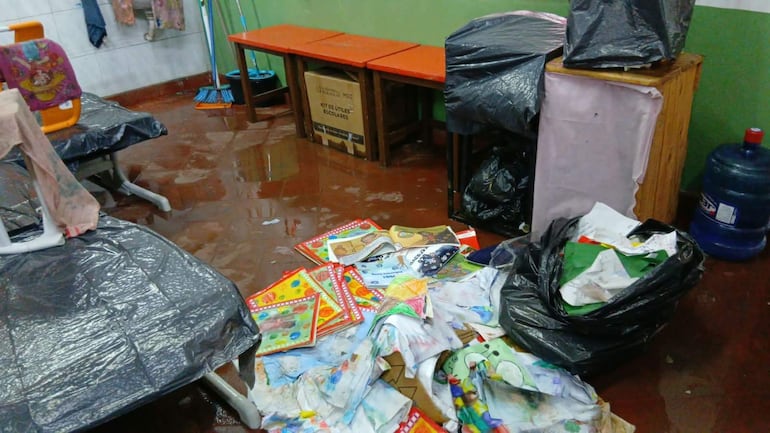 Aula en mal estado con paredes verdes, suelo mojado y papeles esparcidos. Sin personas visibles.
