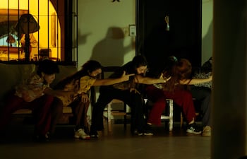 Cinco mujeres sentadas, con expresiones intensas y gestos extendidos, iluminadas suavemente en un entorno acogedor.