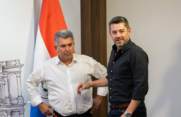 Piris con camisa blanca, pensativo, y Alliana con barba y camisa negra, interactuando en oficina con bandera paraguaya de fondo.