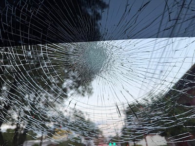 Cristal de vehículo roto por granizo, con patrón de fractura radial en un ambiente urbano con árboles y un semáforo.