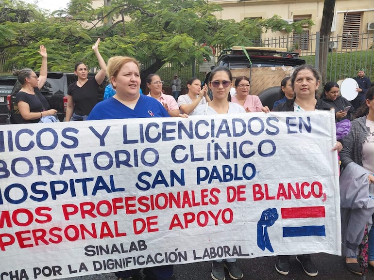 Mujer con blusa azul sostiene pancarta en protesta frente al Hospital San Pablo, rodeada por otras mujeres con carteles.