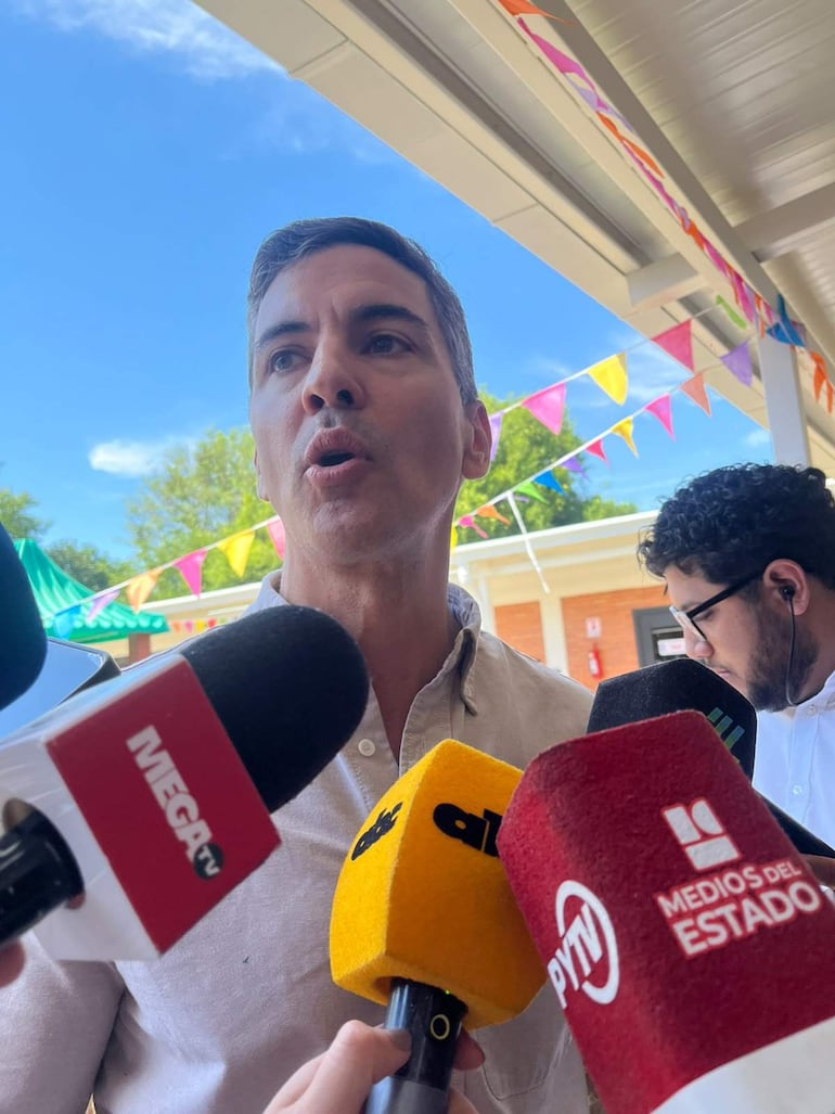 Santiago Peña, serio, frente a una multitud de micrófonos, con un segundo hombre tras él, en un entorno festivo al aire libre.