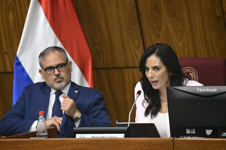 Claudia Centurión con blusa blanca hablando desde un micrófono, mientras un hombre con gafas escucha atentamente en un entorno formal.