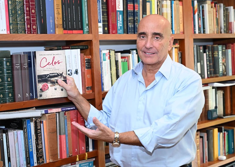 Carlos Mateo Balmelli, con camisa celeste, sostiene el libro 'Celos' en una biblioteca iluminada entre estantes llenos de libros.