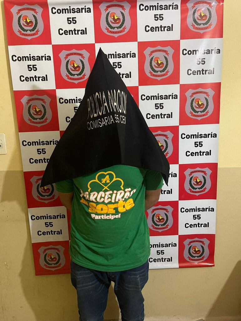 Hombre de espaldas con camiseta verde 'PARCEIRÃO A SORTE' y capucho negro de Policía Nacional en una comisaría.