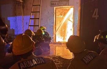 Cuatro bomberos con cascos amarillos observan fuego intenso, preparados con una manguera en un entorno oscuro.