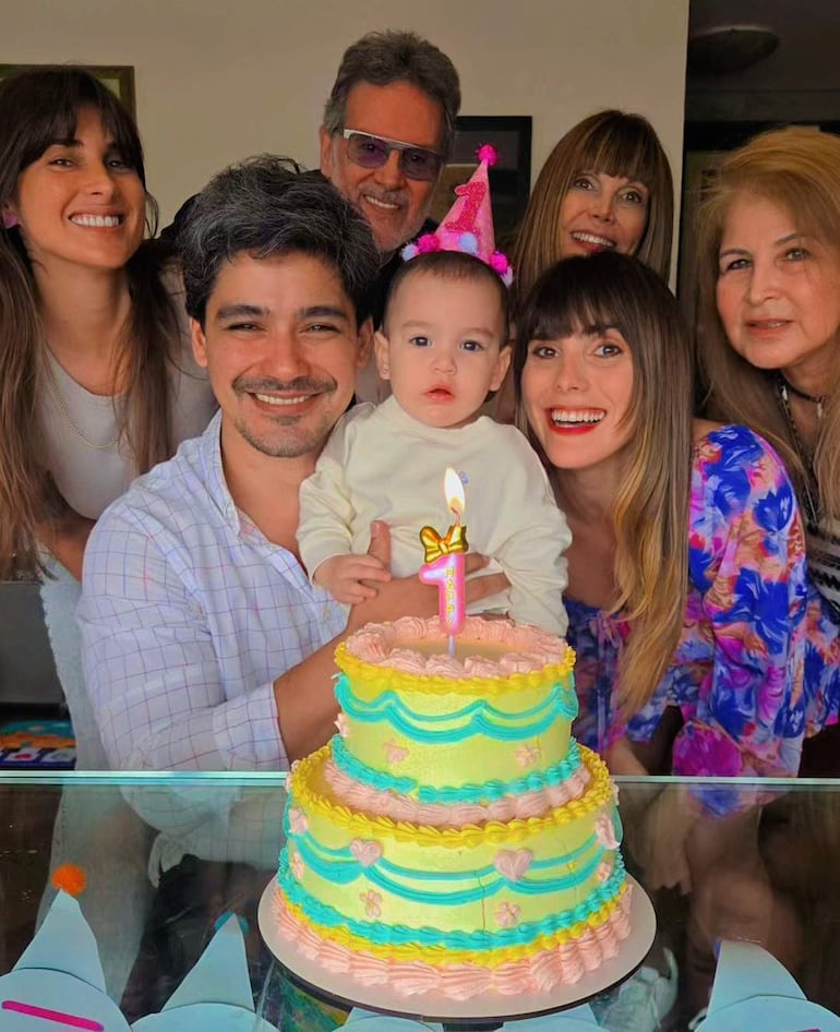 Pelusa Rubin sonríe mientras sostiene a un bebé con gorro de cumpleaños. Amigos felices rodean una mesa con pastel de colores.