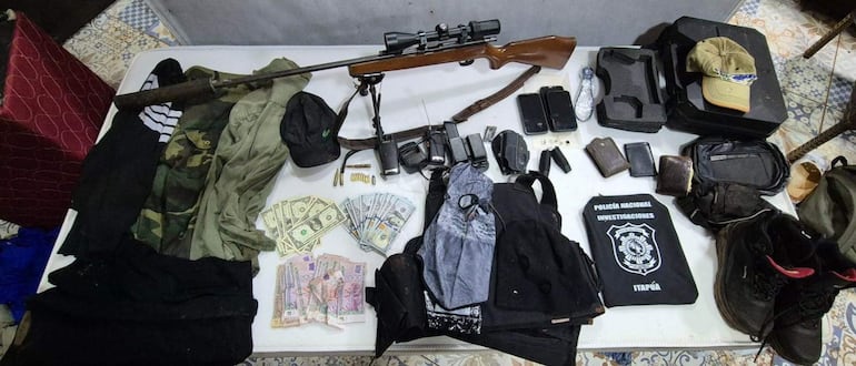 Escena de incautación con un rifle, teléfonos y dinero sobre una mesa en espacio interior, sin personas visibles.