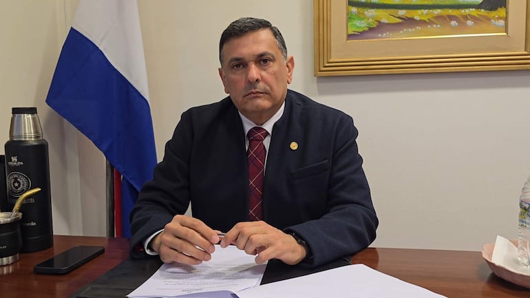 Edgar Olmedo en chaqueta oscura y corbata de cuadros, sentado frente a documentos y una botella de agua en un ambiente de oficina.