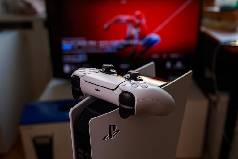 Consola PlayStation 5 blanca con control sobre mesa, televisor muestra juego de Spider-Man en pantalla roja.