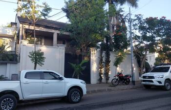 Escena exterior con vehículos, una camioneta blanca y un coche de policía, rodeados de vegetación en una calle tranquila.