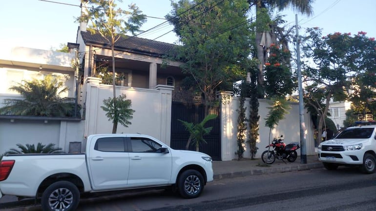 Escena exterior con vehículos, una camioneta blanca y un coche de policía, rodeados de vegetación en una calle tranquila.