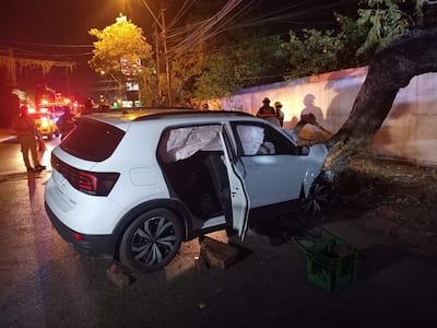 Vehículo blanco dañado contra un árbol, con personal de emergencias en uniforme atendiendo la situación en un ambiente urbano nocturno.
