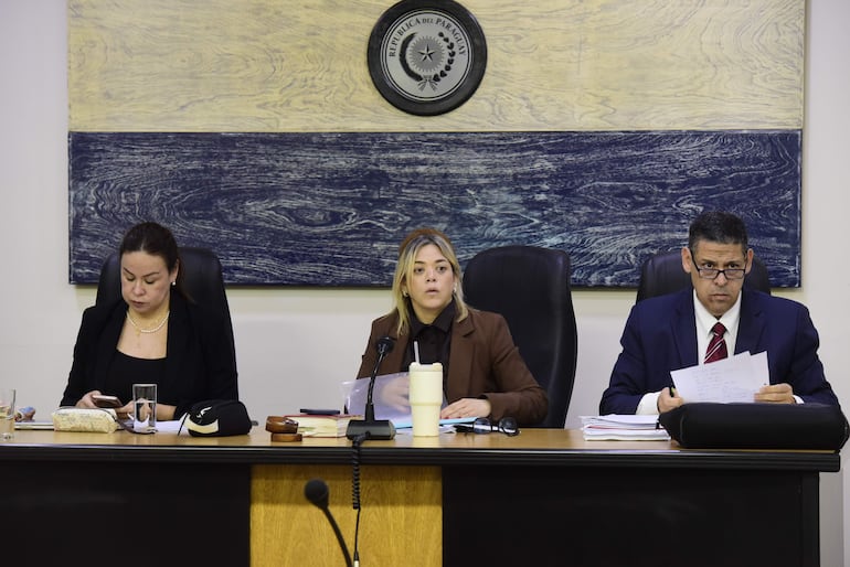 Tres personas en mesa: mujer de negro con teléfono, mujer rubia hablando y hombre en traje con gafas sosteniendo documentos.