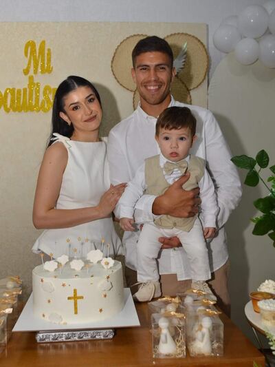 Mujer con blusa blanca y hombre con camisa blanca sonríen, sosteniendo a un niño en un ambiente decorado con globos y un pastel.