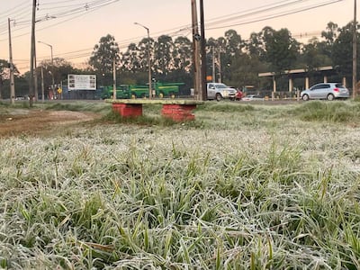 Paisaje urbano frío con césped cubierto de escarcha, vehículos y máquinas agrícolas en el fondo.