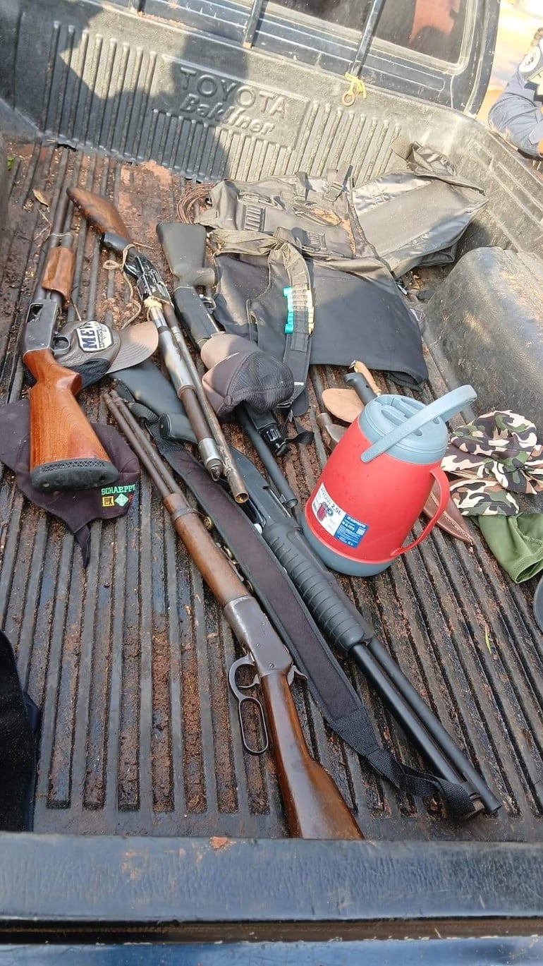 Interior de camioneta Toyota con rifles desordenados, termo rojo y mochila negra en ambiente exterior.