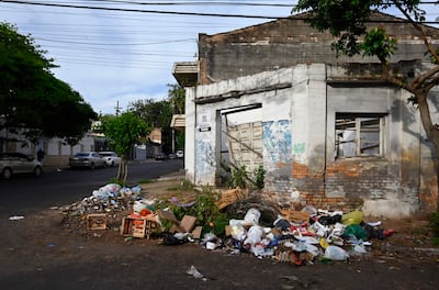 Montón de basura en calle residencial, con edificios deteriorados y vehículos estacionados al fondo.