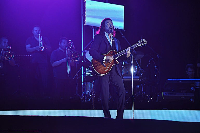 Marco Antonio Solís con guitarra eléctrica y micrófono, vestido formalmente, rodeado de músicos en un escenario iluminado.