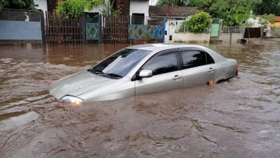 Vehículo sedán plateado sumergido en agua marrón, rodeado de casas y vegetación húmeda tras lluvias recientes.
