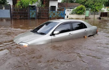 Vehículo sedán plateado sumergido en agua marrón, rodeado de casas y vegetación húmeda tras lluvias recientes.