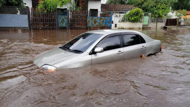 Vehículo sedán plateado sumergido en agua marrón, rodeado de casas y vegetación húmeda tras lluvias recientes.