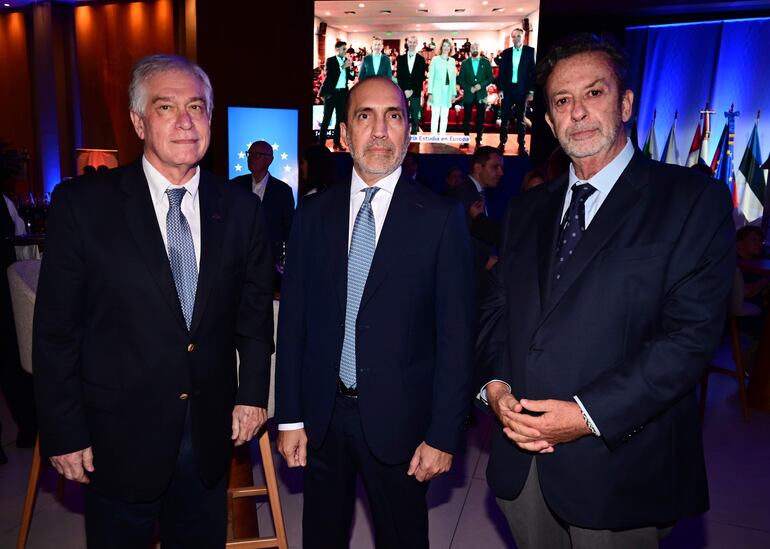 Tres hombres en trajes oscuros, uno con cabello canoso, otro contemplativo y el tercero serio, en un ambiente decorado con luces azules.