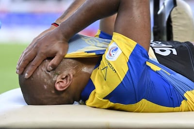 Jugador de fútbol con camiseta azul y amarilla, tendido en camilla y sosteniendo la frente con expresión de dolor.