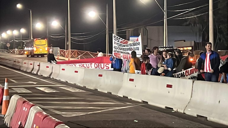 Grupo de personas en protesta con pancartas en un camino; algunos visten camisetas rojas y blancas del Club Olimpia.