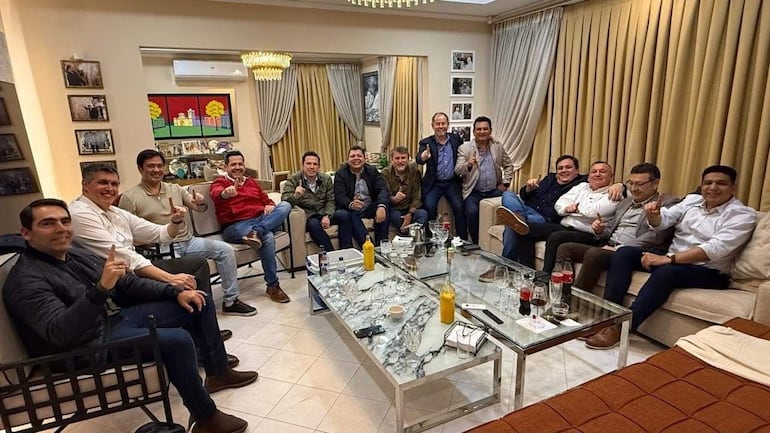 Hugo Meza, Marcelo Soto y otros hombres en un salón conversando alegremente, algunos levantan el dedo índice en un gesto festivo.