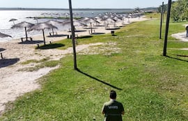 Hombre de espaldas con camiseta 'MADES' caminando sobre césped hacia la playa, sombrillas y bancos de madera en el fondo.