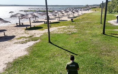 Hombre de espaldas con camiseta 'MADES' caminando sobre césped hacia la playa, sombrillas y bancos de madera en el fondo.