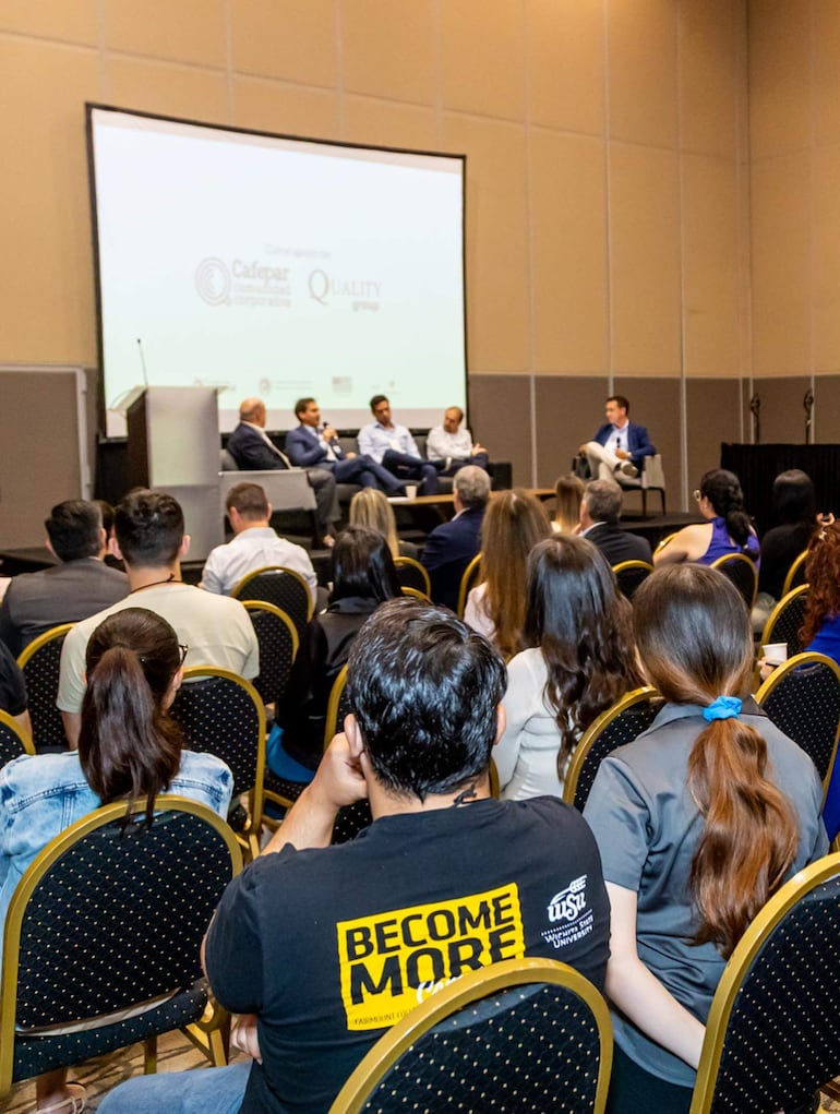 Cinco panelistas sentados, uno hablando mientras el público escucha, todos con camisetas 'BECOME MORE'.