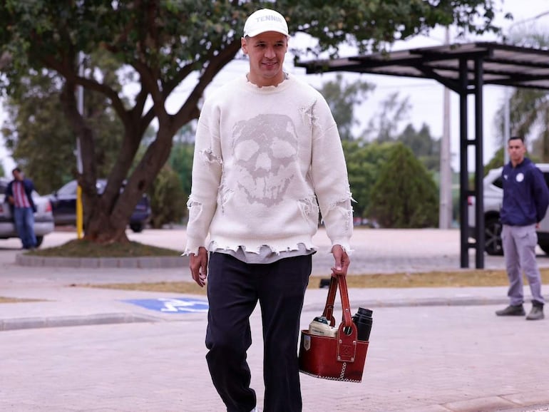 Alex Arce, con suéter claro y pantalones oscuros, lleva un bolso mientras camina. Otro individuo observa al fondo.