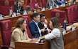 Cristina Villalba en chaqueta beige y otra mujer en blusa blanca conversan, rodeadas de hombres con dispositivos en salón legislativo.