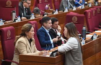 Cristina Villalba en chaqueta beige y otra mujer en blusa blanca conversan, rodeadas de hombres con dispositivos en salón legislativo.