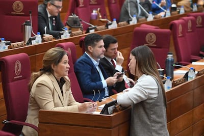 Cristina Villalba en chaqueta beige y otra mujer en blusa blanca conversan, rodeadas de hombres con dispositivos en salón legislativo.