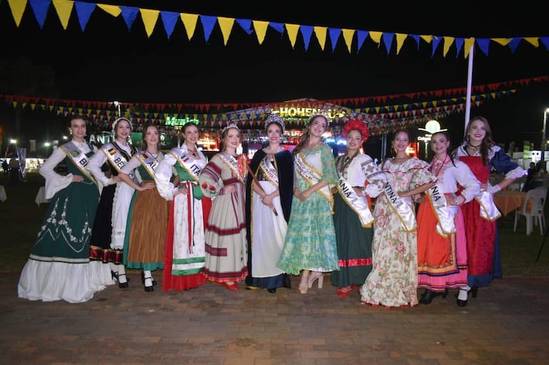 Mujeres sonrientes en trajes tradicionales alineadas en el evento, con cintas de países y decoraciones festivas de fondo.