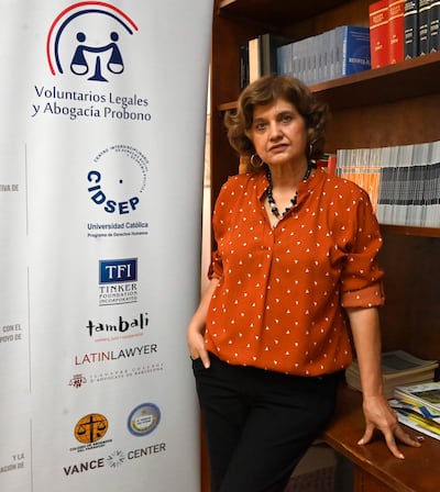 Mónica Cáceres con blusa terracota y collar negro, apoyada en una mesa junto a un panel informativo en una oficina.