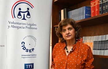 Mónica Cáceres con blusa terracota y collar negro, apoyada en una mesa junto a un panel informativo en una oficina.