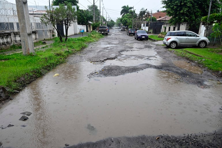 Calle en mal estado con baches y agua acumulada, rodeada de vehículos estacionados y césped verde.