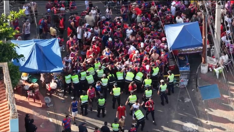 Multitud enérgica con camisetas rojas y azules, policías en chalecos amarillos controlando la situación y carpas azules al fondo.