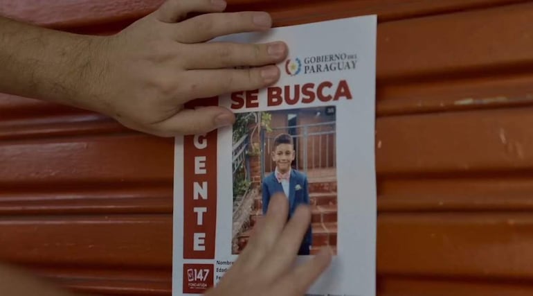 Mano sostiene un cartel rojo que dice 'SE BUSCA', fondo blanco con la foto de un niño sonriendo.