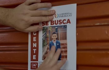 Mano sostiene un cartel rojo que dice 'SE BUSCA', fondo blanco con la foto de un niño sonriendo.