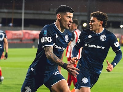 Dos futbolistas con camisetas azules y detalles blancos celebran en el campo, uno apunta al público con gesto entusiasta.